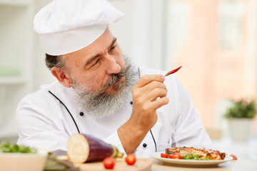 Close up portrait of cheerful senior chef holding red chili pepper while cooking meat in restaurant kitchen, copy space