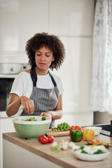 Attractive mixed race woman in apron standing in kitchen and putting chopped lettuce in bowl. Meal preparation concept.