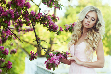 Fototapeta premium Close-up portrait of a beautiful young woman with long blonde hair, perfect skin and beautiful make up in a pink floral summer garden. Tender girl in beige dress like a princess