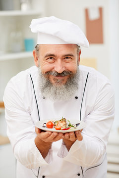 Portrait of bearded senior chef presenting beautiful Italian dish and smiling happily at camera while posing in restaurant kitchen