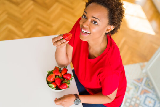 Beautiful young african american woman eating fresh red strawberries at home