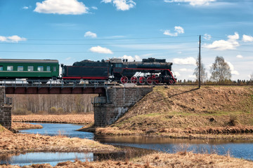 A steam locomotive drives the train over a high embankment over a bridge. A winding river flows under the bridge. Springtime. Railway. Landscape.