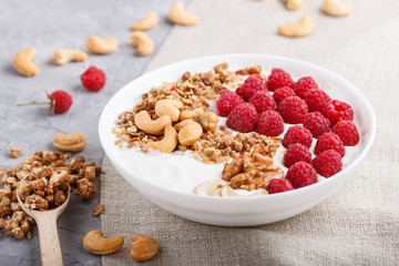 Yoghurt with raspberry, granola, cashew and walnut in white plate with wooden spoon on gray concrete background and linen textile. side view.
