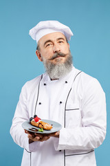 Waist up portrait of cheerful senior chef presenting beautiful dessert while standing against blue background