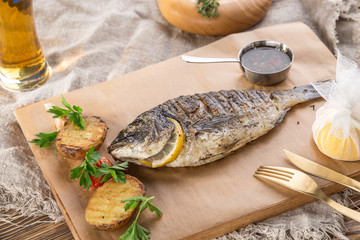 grilled dorado fish with lemon and potato wedges on wooden board and glass of beer on wooden table