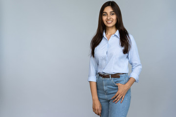 Smiling attractive young adult indian woman in casual wear standing isolated on grey studio background with copy space. Sales professional hindu lady model looking at camera with happy face