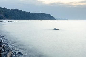 Naklejka na ściany i meble steep coastline on the horizon, smooth surface of the sea at a long exposure with a large stone sticking out of the water in twilight