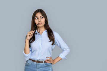 Thoughtful dreamy young adult indian woman looking away thinking doubting or choosing deciding holding glasses in hands standing in casual clothes isolated on grey background with copy space