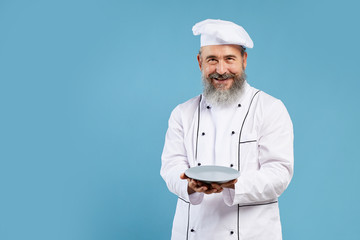 Portrait of bearded senior chef holding empty plate and smiling at camera while standing against blue background, mock up template with copy space