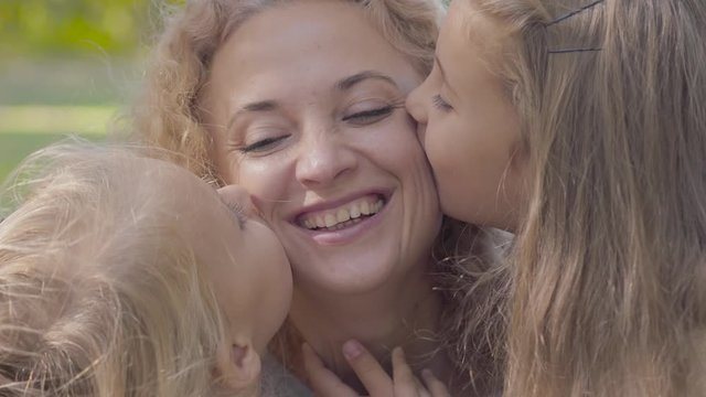 Close-up portrait of two little pretty caucasian girls kissing their mother on cheeks. Happy sisters expressing their love to the smiling woman with blond curly hair.