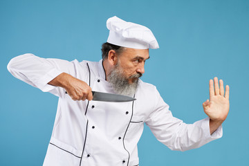 Side view portrait of charismatic bearded chef holding sharp knife standing en garde while posing against blue background in studio, copy space