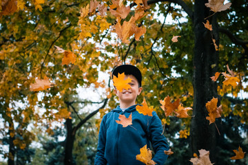 Boy wearing a blue hoody and a black snapback jumps and runs in the autumn park among the flying yellow, orange and red leaves. Colorful forest and seasonal park concept.