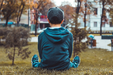 Boy sitting on the grass wearing blue hoody, jeans, black snapback and sneakers in the autumn park....