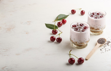 Yoghurt with cherries, chia seeds and granola in glass with wooden spoon on white wooden background. side  view.