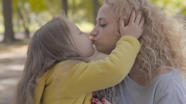 Side view of two little cute caucasian sisters giving their mom flowers and kissing her. Pretty blond curly woman enjoying holidays with her lovely daughters in the autumn park.