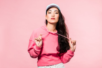 attractive asian woman in pink pullover and cap looking at camera isolated on pink