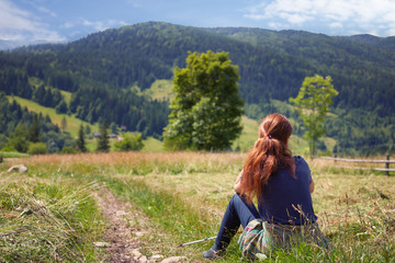 Fototapeta na wymiar Redhead woman hiker in a dark blue t-shirt sitting on a meadow, admiring a mountain view looking out over distant ranges and fir-tree forest. Travel, healthy lifestyle and Relax Concept
