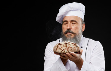 Portrait of charismatic bearded baker holding fresh bread loaf lovingly while standing against black background, copy space - obrazy, fototapety, plakaty