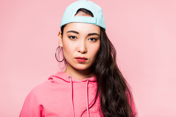 attractive asian woman in pink pullover and cap looking at camera isolated on pink