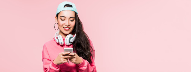 panoramic shot of asian woman in pullover and cap with headphones holding smartphone isolated on pink
