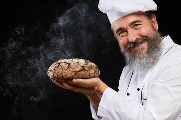 Portrait of charismatic bearded baker holding fresh bread loaf while standing against black...