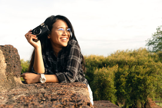 Happy fun smiling Asian woman holding film camera watching summer sunset with warm sun - Millennial girl leaning on brick wall enjoying life - Travel lifestyle blogger taking pictures at golden hour