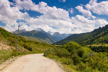Fototapeta na wymiar Country road high in the mountains. Tall trees, snowy mountains and white clouds on a blue sky. Kyrgyzstan Beautiful landscape.