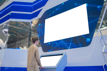 Woman using electronic kiosk and looking at white blank large interactive wall display in dark room...