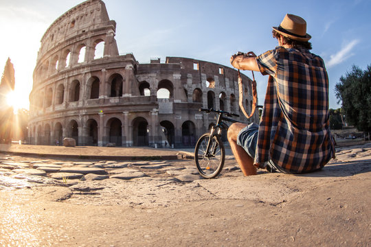 Happy young man tourist wearing shirt and hat with bike taking pictures with vintage camera at colosseum in Rome, Italy at sunrise.