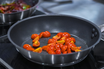 Process of cooking red sliced cherry tomatoes in black frying pan with oil on electric stove at cuisine of restaurant. Professional cooking, catering, culinary, gastronomy and food concept