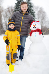 Fototapeta na wymiar Little boy with his father building snowman in snowy park. Active outdoors leisure with children in winter.