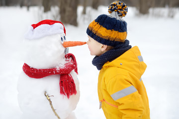 Little boy playing with funny snowman. Child reaches for a snowman's carrot nose and wants to bite....