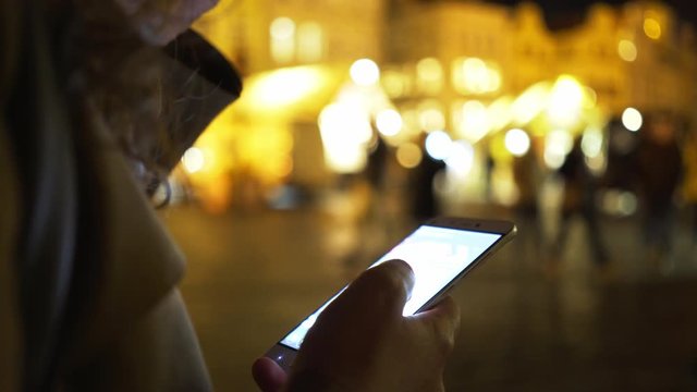Elegant woman in hat at traditional easter fair on main square of Prague in night. Detail shot girl drinking mulled wine and chatting on smartphone at blurred background illuminated street