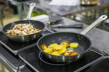 Process of cooking sliced champignon mushrooms and potatoes in frying pans with oil on electric stove at cuisine of restaurant. Professional cooking, catering, cookery, gastronomy and food concept