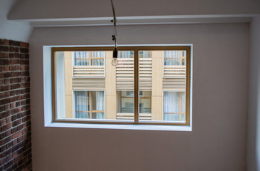 A new empty loft-style apartment, with a brick wall, white walls and a huge window through which you can see the modern new house opposite. One light bulb hangs from the ceiling