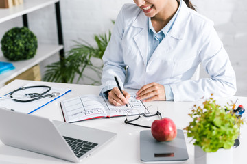 cropped view of happy doctor writing meal plan in notebook