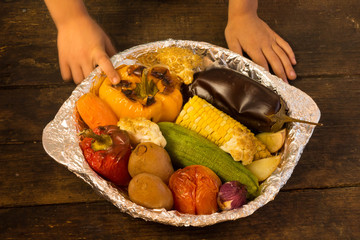  Hand of a child touching baked vegetables in foil potato carrot pepper onion corn on the cob...