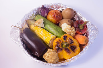 Male hand holding baked vegetables in foil potato carrot pepper onion corn on the cob eggplant cauliflower squash on wooden table