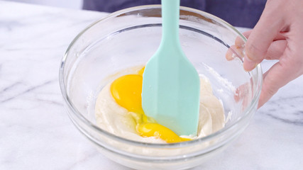 Fototapeta na wymiar Mixing egg yolk into cake batter with green rubber spatula mixer tool stirring until smooth and blend well in a glass bowl, close up, lifestyle