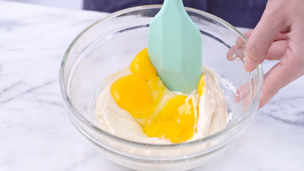 Mixing egg yolk into cake batter with green rubber spatula mixer tool stirring until smooth and blend well in a glass bowl, close up, lifestyle