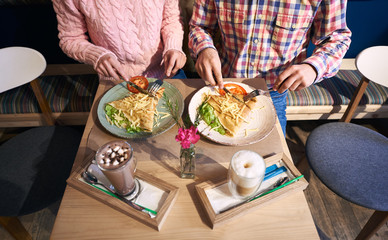 Cropped view of couple - man and woman sitting together in cafe at small table enjoying big pancake sandwich with vegetables. Coffee beverage and vase with flower on blurred foreground. Top view