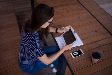 A serious brunette girl, freelancer, with large wrist watch, works at a wooden table on tablet,...