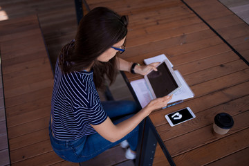 A serious brunette girl, freelancer, with large wrist watch, works at a wooden table on tablet,...