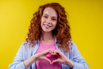 happy young redhaired ginger woman with curly hairstyle pretty face wearing cotton pink shirt ,making heart gesture with hands symbol in studio yellow background