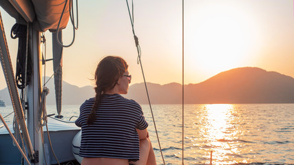 attractive young woman in a striped t-shirt enjoys the sunset on the deck of a sailing yacht....