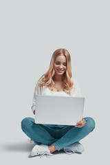 Attractive young woman using laptop and smiling while sitting against grey background