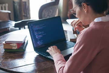 Hacking system. Excited girl with dark hair and in pink sweater and glasses writes code on a laptop in a library.