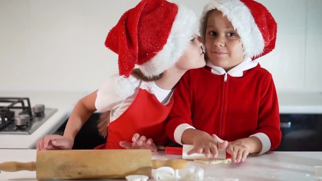 Happy children making Christmas cookies at home, cute little kids wearing red Santa Claus hats, preparing festive sweets. Two cute little sisters kiss each other on New Year's Eve. Slow motion.