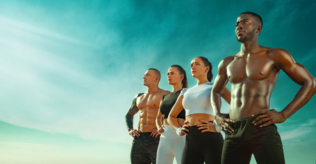 Group of four young sportsmens women and men, fit athletes are standing on the sky background...