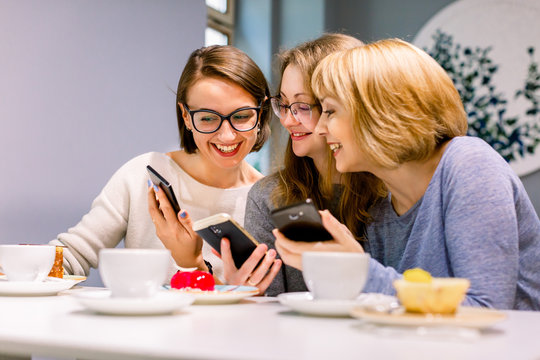 Image of three excited happy pretty girls friends sitting in cafe, drinking coffee, eating cakes and using phones.